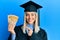 Beautiful blonde woman wearing graduation cap and ceremony robe holding philippine pesos smiling happy pointing with hand and