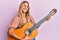 Beautiful blonde young woman playing classical guitar smiling and laughing hard out loud because funny crazy joke