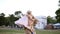 A beautiful blondy girl in a pink dress runs after her mother on the lawn. An adult mother plays with her in the park