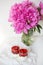 A beautiful bouquet of pink peonies in a vase on a white table stands along with a heart-shaped strawberry dessert. St. Valentine