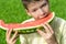 Beautiful boy bites a large slice of red watermelon, on a background of green grass