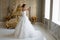 A beautiful bride with chic makeup and hairstyle in a white wedding dress is standing in a bedroom