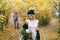Beautiful bride in a knitted hat with a pompon are posing in autumn forest on blurred groom`s background. Wedding
