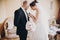 Beautiful bride putting stylish boutonniere on groom suit, standing in room. Gorgeous woman in white gown and man in suit getting