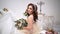 Beautiful bride sits surrounded by wedding dresses, around bouquets and decor. Looks directly at the camera, big