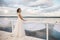 Beautiful bride stands on the pier. Water and cloudy sky on the background