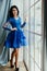 Beautiful brunette in a blue dress at a wide window. A woman in a chic studio interior interior.
