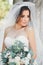 Beautiful brunette bride in elegant white dress holding bouquet posing neat trees