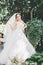 Beautiful brunette bride in elegant white dress holding bouquet posing neat trees