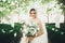 Beautiful brunette bride in elegant white dress holding bouquet posing neat trees