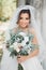 Beautiful brunette bride in elegant white dress holding bouquet posing neat trees