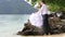 beautiful brunette bride sitting on rock embraces groom