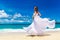 Beautiful brunette bride in white wedding dress with big long white train and with wedding bouquet stand on shore sea. Tropical s