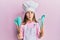 Beautiful brunette little girl wearing professional cook apron holding cooking tools making fish face with mouth and squinting