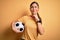 Beautiful brunette player woman playing soccer using football ball over yellow background serious face thinking about question,