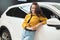 Beautiful brunette woman looking for new car in dealership center leaning on white vehicle looking happy