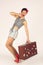 Beautiful brunette woman in pin up style stands and hardly holds a heavy suitcase for travel, white background