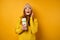 A beautiful brunette in a yellow sweater and hat stands on a yellow background with a white thermos in hands and looks