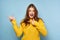 A beautiful brunette in a yellow sweater stands on a blue background, cramping her face and pointing her fingers to the