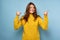 A beautiful brunette in a yellow sweater stands on a blue background, smiling embarrassed, raising her thumbs to the top