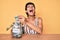 Beautiful brunettte woman holding charity jar with money angry and mad screaming frustrated and furious, shouting with anger