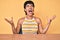 Beautiful brunettte woman sitting on the table over yellow background crazy and mad shouting and yelling with aggressive