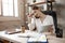 Beautiful busy young woman sit at table in room. She talk on phone and look at notebook. Model hold pen and tablet.