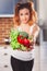 Beautiful caucasian girl holding glasses bowl with ingredients for low-caloric vegetables salad on kitchen.