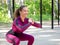 Beautiful caucasian girl in magenta sportswear does squats in sports area outdoor. Workout in the park, in summer