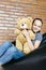 Beautiful caucasian teen girl sitting in black bean bag chair holding big brown teddy bear toy and smiling in camera. Casual outfi