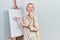 Beautiful caucasian woman with blond hair standing by painter easel stand presenting canva smiling and laughing hard out loud