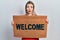 Beautiful caucasian woman holding welcome doormat in shock face, looking skeptical and sarcastic, surprised with open mouth