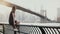 Beautiful Caucasian young woman sitting on river embankment fence at Brooklyn Bridge New York, jetski passing by 4K.