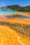 Beautiful cerulean geyser surrounded by colorful layers of bacteria, against cloudy blue sky.