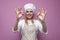 Beautiful cheerful girl cook in uniform and apron on a colored pink background shows okay, portrait of a housewife woman in