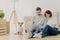 Beautiful couple with dog sit on floor in spacious living room, make purchasing online, buy furniture for new home, have to unpack