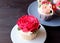 Beautiful cupcake topped with red rose shaped whipped cream with blurry plate of cupcakes in background