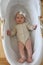 A beautiful cute baby girl in a beige bodysuit with a white bow on her head lies in a white cradle made of light wood.