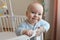 A beautiful cute European baby is standing in a white oval four-poster bed and smiling.