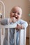 A beautiful cute European baby is standing in a white oval four-poster bed and smiling.