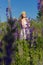Beautiful cute gentle girl in white sundress with a wreath of roses on her head walks a field with lupine