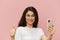 beautiful cute woman with a trendy phone in her hands stands on a pink background in a white t-shirt, holds her thumb up