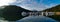 Beautiful early morning panoramic view of a creek with reflections of blue sky, boats, foggy mountains and trees on water