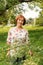 A beautiful elderly woman in flaxen clothes stands tranquilly in her green garden