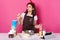 Beautiful European woman spends time at kitchen with whisk in one hand, wearing white t shirt, brown apron and red bright headband