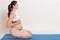 Beautiful expectant mother doing yoga at home, touching her bare belly, looking straight ahead, posing against white background,