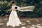 Beautiful fairytale bride in white wedding dress posing with red flowers bouquet while standing on a park road in summer, wind