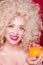 Beautiful fashionable blonde girl in retro style with voluminous curly hairstyle, bare shoulders and an orange in her hands looks