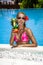 Beautiful female model in pink bikini is standing in the pool and holding colorful fruity drink. Sexy woman in sequin bikini is