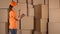 Beautiful female warehouse worker in orange uniform counting items and making records against brown cartons backround